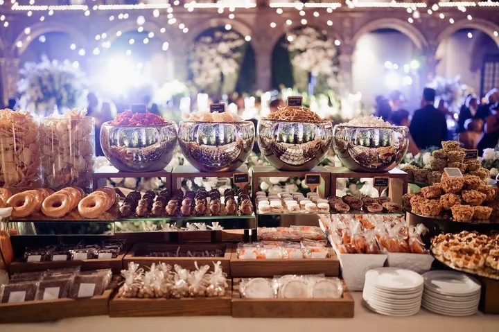 Buffet de postres de boda con donuts, pasteles y cuencos plateados bajo guirnaldas de luces