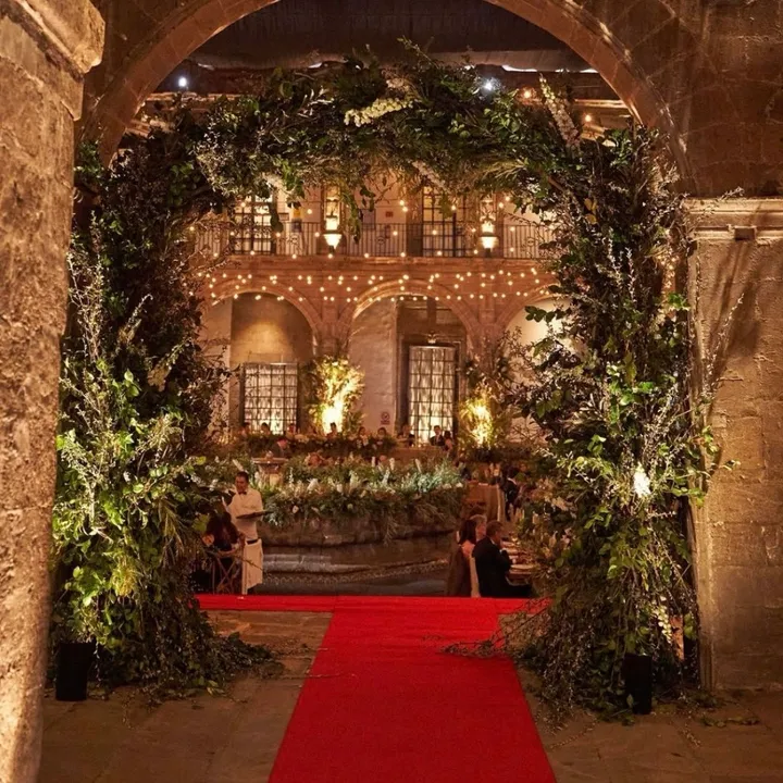 Arco cubierto de vegetación sobre una alfombra roja que da a un patio histórico iluminado con velas