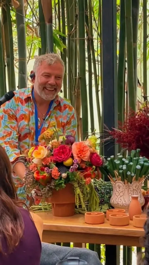 Diseñador floral sonriendo junto a un arreglo de flores de colores en una maceta de barro en un taller
