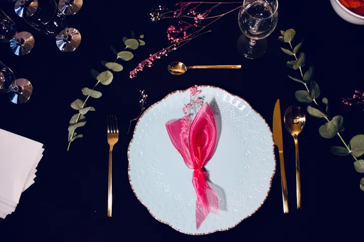 Montaje de mesa elegante con servilleta rosa en forma de lazo, cubertería dorada y eucalipto sobre mesa oscura