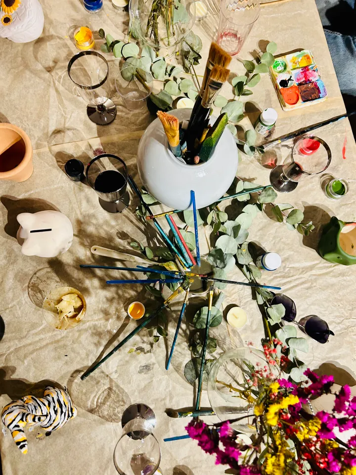 Vista cenital de una mesa de taller con pinceles en un cuenco, eucalipto, pinturas y copas de vino