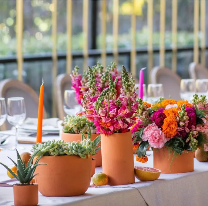 Mesa de boda con macetas de barro con flores rosas y suculentas y velas naranjas junto a una ventana