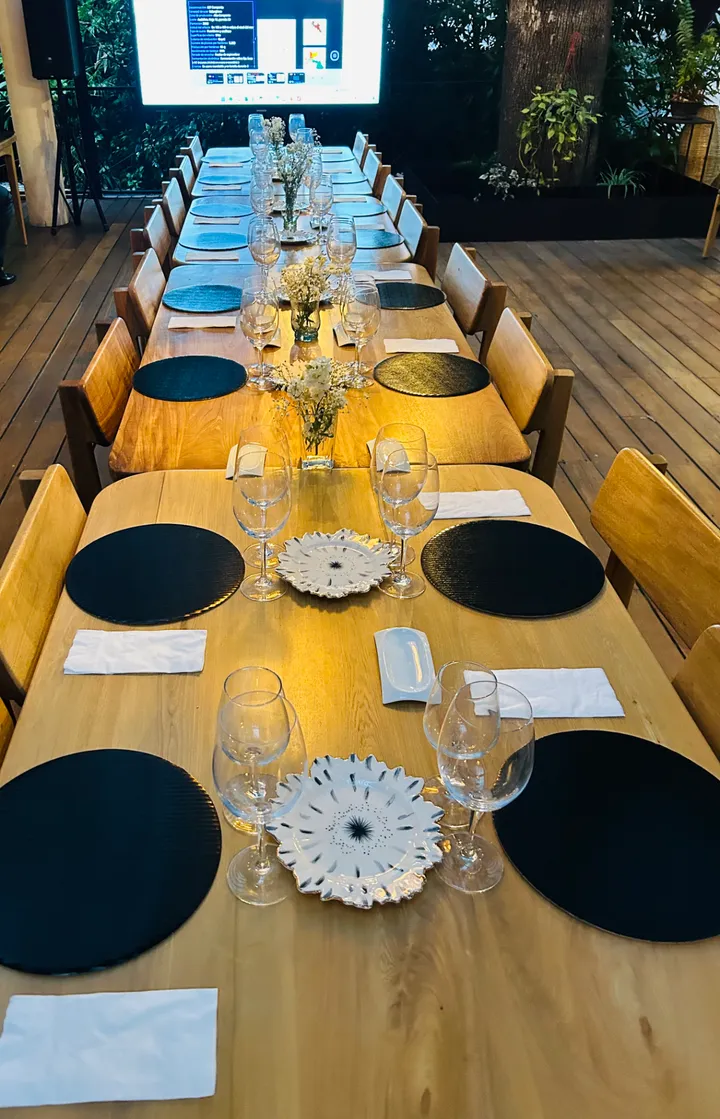 Larga mesa de banquete con bajoplatos negros y copas, y una pantalla de proyección en la cabecera
