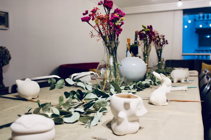 Mesa de taller de pintura en cerámica con figuras blancas sin pintar, flores secas y un camino de eucalipto