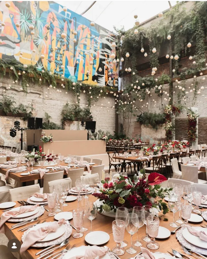 Banquete de boda en nave industrial con un mural colorido, vegetación colgante y mesas con flores rojas