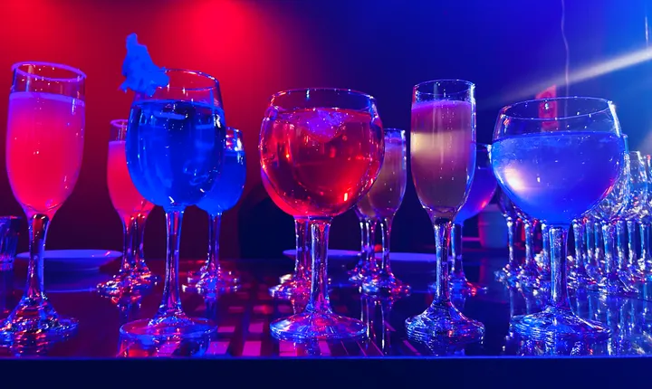Row of colourful cocktails in glasses under dramatic red and blue lighting on a reflective bar